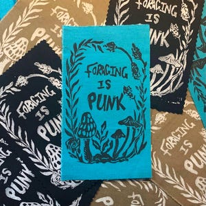 May include: A collection of fabric patches with a hand-printed design. The central patch is turquoise with the text "FORAGING IS PUNK" surrounded by a botanical illustration. Other patches are black and tan with similar designs.