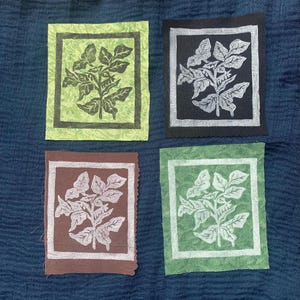 May include: Four handmade paper art pieces with botanical designs. Each square features a plant illustration in white against backgrounds of green, black, brown, and green. The art pieces have a textured appearance and are arranged on a blue fabric.