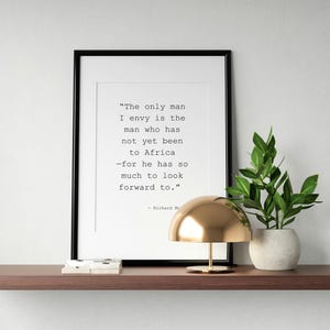 May include: A black framed print with a quote by Richard M. about the man who has not yet been to Africa. The print is on a shelf with a gold lamp and a potted plant.