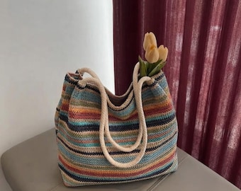 May include: A woven tote bag featuring horizontal stripes in shades of blue, orange, yellow, and brown. The bag has two thick, cream-colored rope handles and contains a small bouquet of pale yellow tulip flowers emerging from the top opening.
