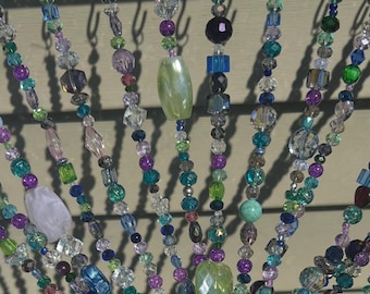 Op de afbeelding: Een kralengordijn met strengen van verschillende maten en vormen kralen in tinten blauw, groen, paars en helder. Het gordijn wordt in een hand gehouden, waarbij het zonlicht schaduwen op de muur erachter werpt.