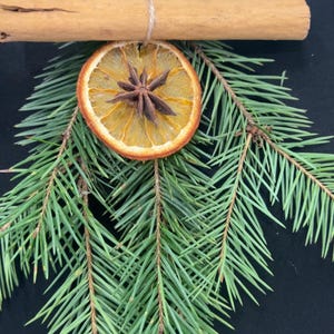May include: A festive arrangement featuring a cinnamon stick, dried orange slice, and star anise, tied together with twine and set atop fresh green pine branches. This decorative piece evokes a natural, rustic aesthetic, perfect for holiday decor.