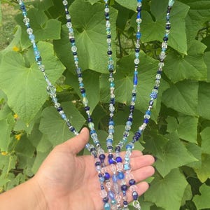 May include: A beaded hanging ornament with multiple strands. The strands are adorned with a variety of blue and clear beads, including faceted crystals and small, round beads. The ornament is suspended from a metal ring, creating a decorative effect.
