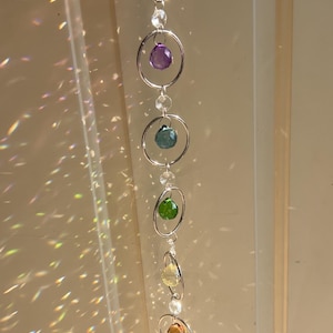 May include: A hanging crystal suncatcher with seven colorful gemstones. Each gem is set within a silver-tone ring, suspended from a chain. The gemstones are red, purple, blue, green, yellow, and orange.