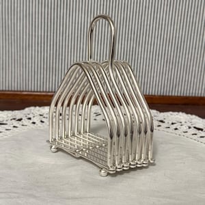 May include: A silver-colored, vintage-style toast rack. The rack has a rectangular base with rounded feet and a handle. The design features multiple vertical bars to hold toast slices. The background is a white cloth with a lace trim.