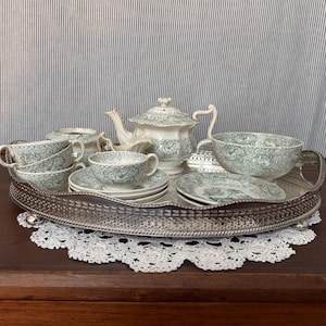 May include: A vintage tea set with a green and white floral pattern, including a teapot, sugar bowl, creamer, cups, saucers, and a large serving bowl. The set is arranged on a silver tray with a lace doily underneath.