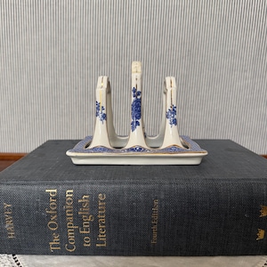 May include: A white ceramic toast rack featuring blue floral patterns and gold trim along the edges. The rack has three vertical dividers and a rectangular base, resting on a dark gray book titled The Oxford Companion to English Literature.