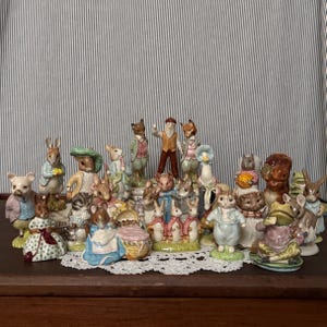 May include: A large collection of hand-painted ceramic animal figurines inspired by Beatrix Potter characters. The group includes various rabbits, a pig, a frog, a fox, and other woodland creatures dressed in colorful Victorian-style clothing.