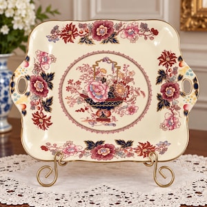 May include: A square ceramic serving tray with rounded corners and side handles, featuring a cream-colored base with a central floral basket motif and a border of red, pink, and dark blue flowers, finished with a gold-toned rim.