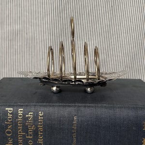 May include: Silver-toned metal toast rack with six arched slots and a decorative, scalloped base. The rack is resting on a dark blue book titled "The Oxford Companion to English Literature, Fourth Edition."