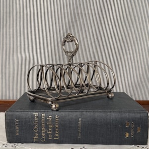 May include: A silver-toned metal toast rack with a decorative handle and six slots. The rack sits atop a dark blue book titled "The Oxford Companion to English Literature." The background features a striped pattern.