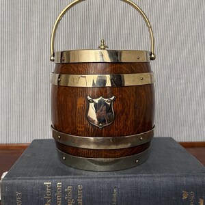 May include: A barrel-shaped ice bucket made of dark brown wood with polished brass bands, a brass handle, and a central brass shield emblem. The lid features a small brass knob. The bucket is resting on a large, thick, gray hardcover book.