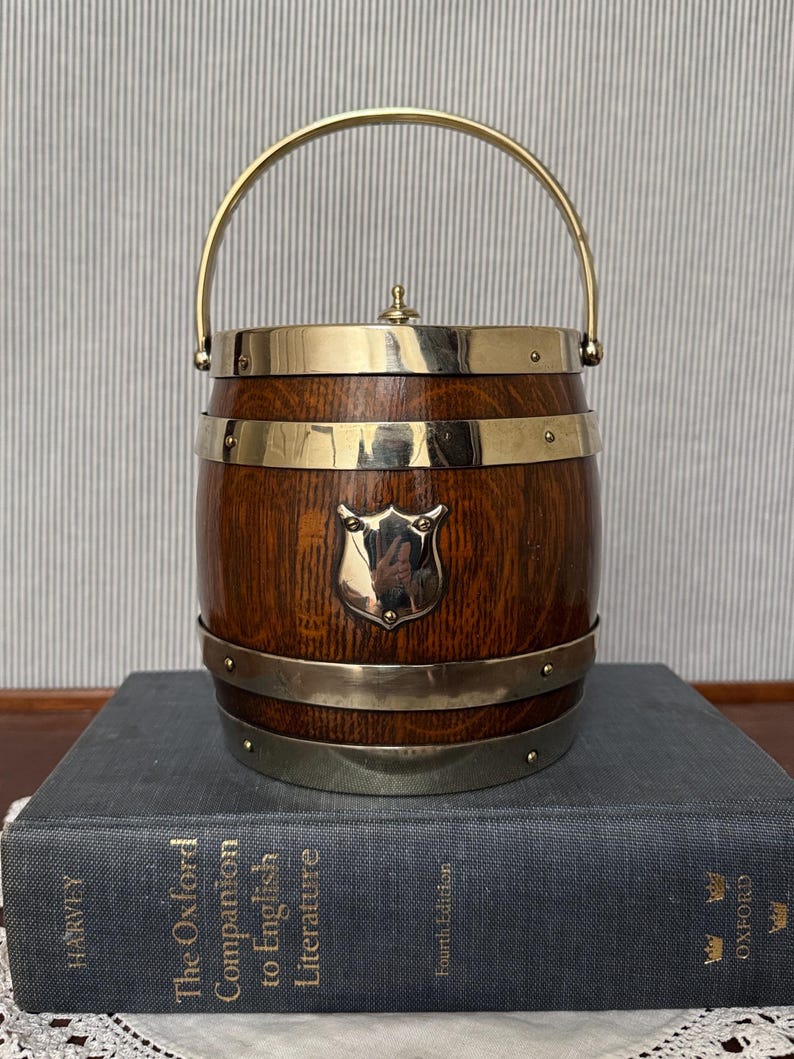 May include: A barrel-shaped ice bucket made of dark brown wood with polished brass bands, a brass handle, and a central brass shield emblem. The lid features a small brass knob. The bucket is resting on a large, thick, gray hardcover book.