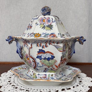 May include: A vintage, ornate soup tureen with a matching lid and base. The tureen features a floral and bird design in shades of blue, orange, pink, and green. The handles and finial are accented with gold. The tureen sits on a white lace doily.