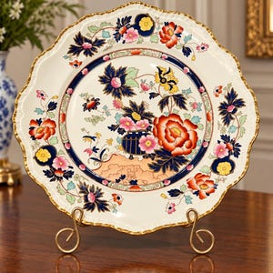 May include: A round ceramic decorative plate with a scalloped edge and gold trim. The center features a colorful floral pattern with red, pink, yellow, and dark blue flowers on a cream background, set within a dark blue circular border.