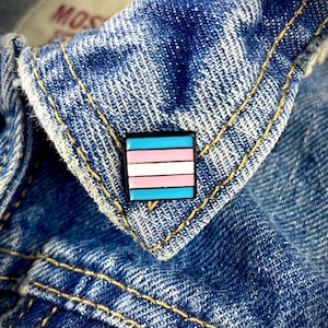May include: A blue denim jacket with a small, square transgender pride pin. The pin has five horizontal stripes: light blue, pink, white, pink, and light blue.