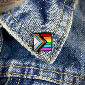 Puede incluir: Un pin cuadrado con contorno negro con un diseño de bandera arcoíris. La bandera tiene un triángulo negro en el centro con una franja blanca a la izquierda, una franja azul claro a la derecha y una franja rosa en la parte inferior. Las franjas arcoíris están en la parte superior del pin.