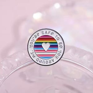 May include: White enamel pin with a rainbow and white stripes design. The pin says "You're safe to be you around me" with a white heart in the centre.