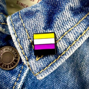 May include: A yellow, white, and purple striped pin with a black border. The pin is on a blue denim jacket.