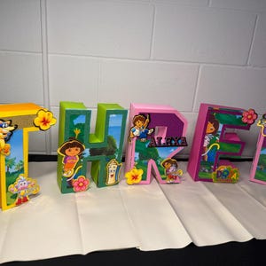 Könnte beinhalten: Bunte, dekorative Buchstaben, die den Namen "Alexa" buchstabieren. Die Buchstaben sind gelb, grün und rosa, verziert mit Dora the Explorer-Charakteren und floralen Akzenten. Der Buchstabe "R" trägt den Namen "Alexa".