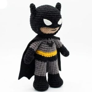 May include: A handmade crochet doll of a superhero character featuring a black cowl with pointed ears, white eye patches, and a tan face. The figure wears a grey suit with a yellow belt, a black chest emblem, and a black cape attached to the back.