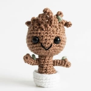 May include: A brown crocheted amigurumi figure with a large head, two black plastic eyes, and a stitched smile. The figure has small green leaf accents on its head and arms, and it is standing inside a small, white crocheted pot base.