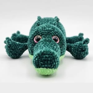 May include: A handmade, plush green crocodile toy with a light green snout and large, pink-rimmed eyes. The crocodile is lying down, with its arms and legs outstretched. The toy is made of a soft, textured material.