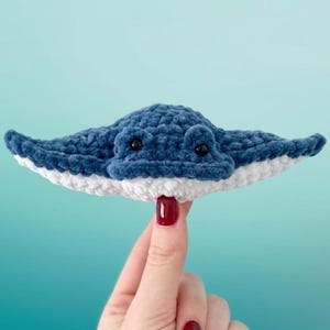 Può includere: Una manta fatta a mano all'uncinetto, blu e bianca, con occhi di sicurezza neri. Il peluche è tenuto in mano su uno sfondo azzurro. La manta ha un aspetto morbido e strutturato.