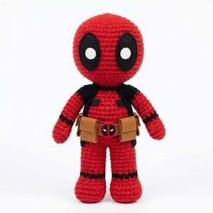 May include: A crochet doll featuring a red and black color scheme with a large round head, black eye patches with white circular centers, and a tan utility belt around the waist that includes a small circular buckle with a red and black logo.