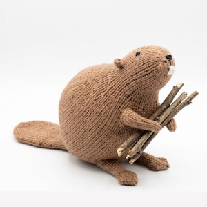 May include: A brown knitted beaver stuffed animal with a flat tail and small ears. The beaver has black eyes, a small black nose, and two white teeth. It is holding a bundle of three thin, brown wooden sticks in its front paws.