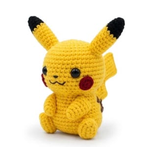May include: A yellow crochet plush toy shaped like a creature with long ears tipped in black, round black eyes, a small black nose, a stitched mouth, and circular red patches on the cheeks. The figure has a lightning bolt shaped tail and brown markings.