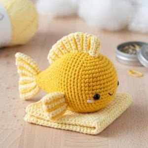 Könnte beinhalten: Ein handgefertigter, gelber Häkelfisch mit hellgelben Flossen und Schwanz, der auf einem passenden gelben gehäkelten Quadrat ruht. Der Fisch hat schwarze Augen und einen kleinen rosa Mund. Das Bastelprojekt ist ein niedliches, skurriles Design.