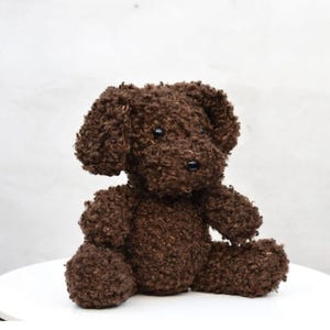 May include: A brown plush dog toy with a curly texture. The stuffed animal has black button eyes and a black nose. The dog is sitting on a white surface.