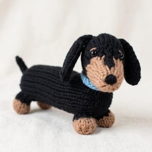 Puede incluir: Un juguete de perro salchicha tejido en negro y marrón claro con un collar azul. El juguete tiene un cuerpo largo, orejas caídas y patas pequeñas. El perro está de pie sobre sus cuatro patas.