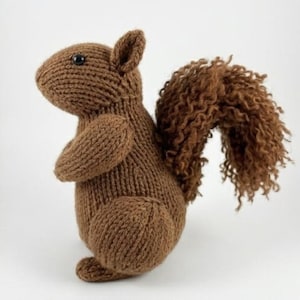 May include: A brown knitted squirrel toy featuring a smooth, stockinette-stitched body and a large, textured, curly-knit tail. The squirrel has a small, dark, round eye and is positioned in a seated posture against a plain white background.