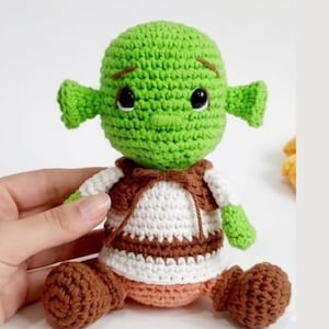 May include: A handmade crochet doll of a green ogre character with large ears. The doll wears a white shirt with a brown vest and brown pants. The doll has black eyes and a small smile. The doll is a whimsical, handcrafted item.