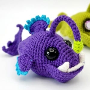 May include: A handmade purple crochet anglerfish with blue and green accents. The fish has large black eyes, white teeth, and a glowing lure. The anglerfish is a whimsical, soft toy.