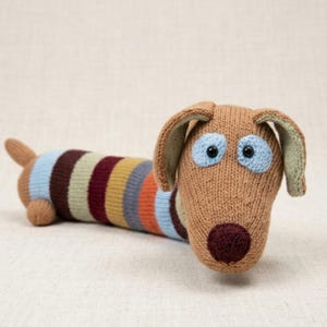 May include: A knitted dachshund dog toy with a brown head, ears, and tail. The body is striped with blue, brown, orange, and gray. The dog has blue eyes and a dark brown nose. The toy is made of soft yarn.