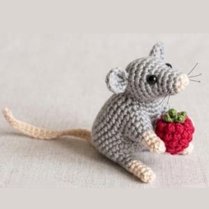 Puede incluir: Un pequeño ratón de ganchillo gris con ojos negros y bigotes, que sostiene una frambuesa de ganchillo roja brillante y texturizada con un tallo verde. El ratón tiene patas de color beige claro y una cola larga, fina y beige claro.