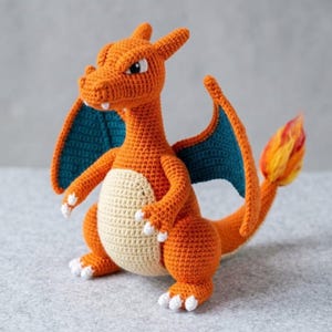 May include: A crochet plush toy shaped like a dragon with orange skin, a cream-colored belly, and teal-colored wings. The creature has white claws, small white teeth, and a tail ending in a flame-like tuft of yellow and orange yarn fibers.
