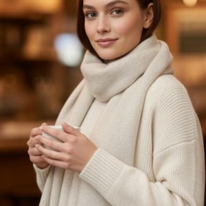 May include: A woman wearing a cream-colored sweater and scarf, holding a white mug. The scarf has a fringed edge. The background is softly blurred with warm tones, suggesting an indoor setting. The sweater has a ribbed knit design.