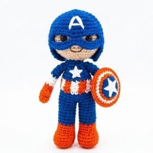 May include: A handmade crochet doll of a superhero character wearing a blue mask with a white letter A, a blue top with a white star, and red and white striped midsection. The doll has orange boots and holds a round red, white, and blue shield.