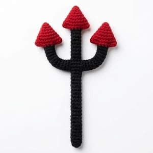 May include: A crochet trident toy featuring a long black handle that splits into three prongs. Each of the three prongs is topped with a small, textured red triangular point. The entire object is crafted from yarn with a visible stitched pattern.
