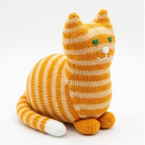 Puede incluir: Un juguete de gato tejido con rayas horizontales de color naranja y crema. El gato tiene ojos verdes bordados, una nariz blanca y una boca blanca cosida. La cola es a rayas con una punta blanca sólida y el cuerpo está sentado.