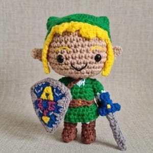 Peut inclure: Une poupée amigurumi crochetée représentant un personnage aux cheveux jaunes, portant un bonnet vert et une tunique verte. La poupée tient un bouclier bleu et gris avec un symbole jaune et une épée bleue et grise. Yeux noirs et oreilles pointues.