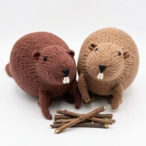 May include: Two round, knitted beaver plush toys, one in dark brown and one in light tan, each featuring small black eyes, a black stitched nose, and two white teeth, positioned behind a small pile of thin, natural wooden sticks.