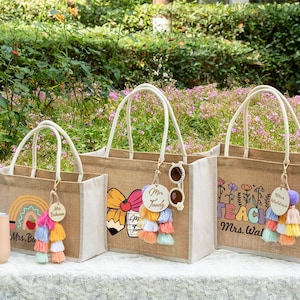 May include: Three rectangular jute tote bags with white rope handles and personalized wooden tags. The bags feature colorful graphic prints including a rainbow with an apple, a pencil with a bow, and floral designs with the text TEACH Mrs. Waldron.