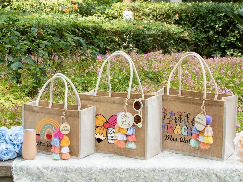 May include: Three rectangular jute tote bags with white rope handles and personalized wooden tags. The bags feature colorful graphic prints including a rainbow with an apple, a pencil with a bow, and floral designs with the text TEACH Mrs. Waldron.