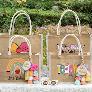 May include: Four rectangular jute tote bags with white handles, featuring colorful graphic prints of rainbows, apples, and school supplies. Two bags include personalized wooden tags labeled Mrs. Balaam and Mrs. Helena with multi-colored tassels.