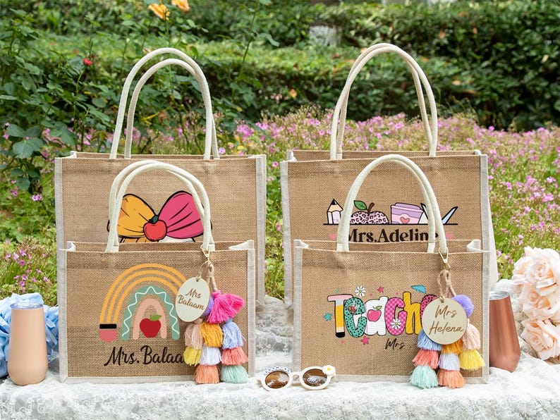 May include: Four rectangular jute tote bags with white handles, featuring colorful graphic prints of rainbows, apples, and school supplies. Two bags include personalized wooden tags labeled Mrs. Balaam and Mrs. Helena with multi-colored tassels.